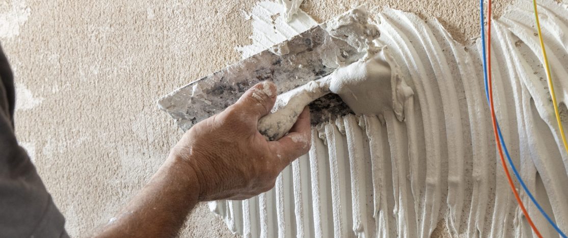 Craftsman spreads tile adhesive with notched trowel on a untiled house wall surface, tiler hand closeup. New building construction or renovation.